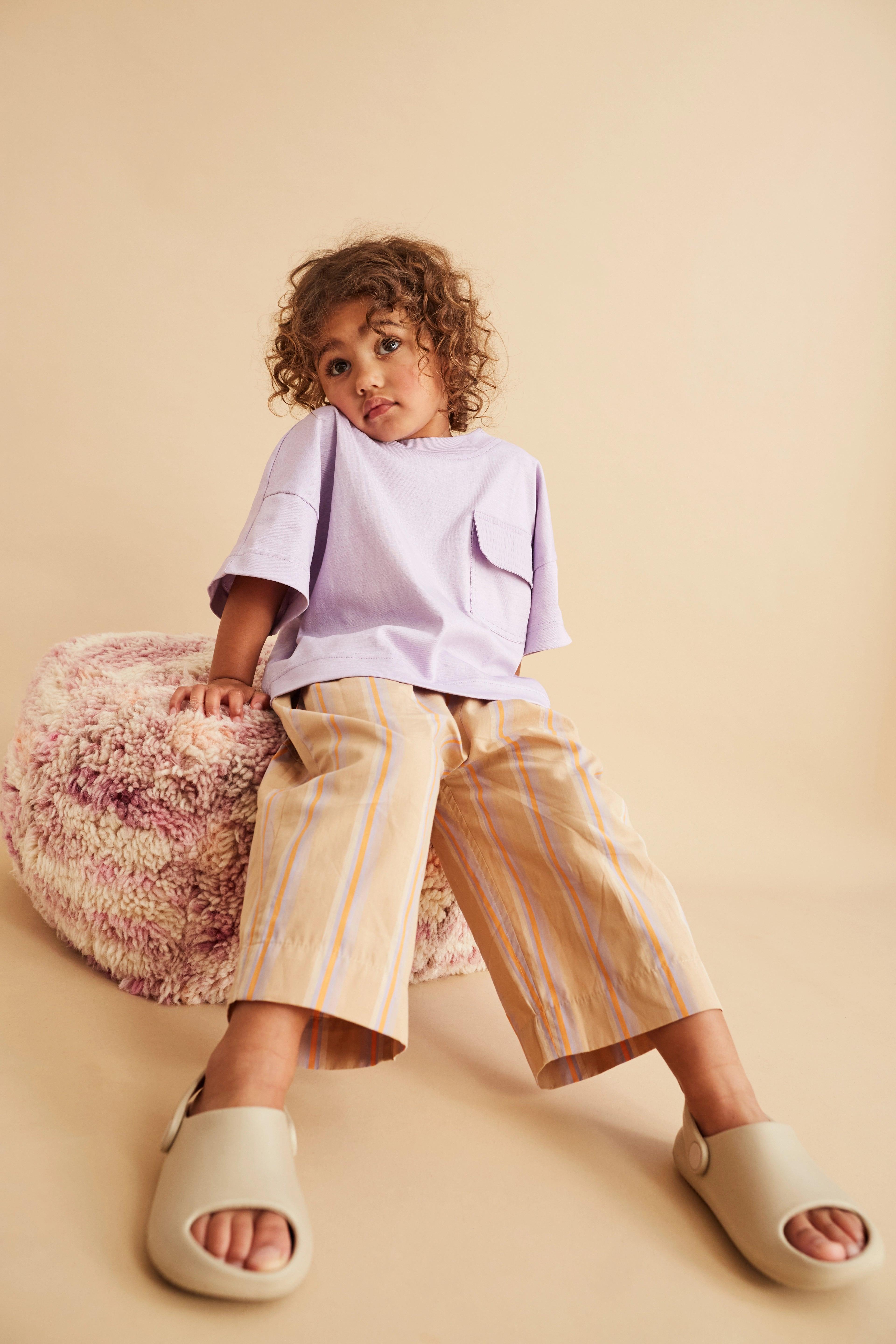 Modern and comfy straight leg striped trousers with elasticated waistband and slanted side pockets, patch pocket in the back. Made from a soft, washed organic cotton, beige base with lilac and orange yarn-dyed stripe. Matching woven t-shirt and romper available for the perfect kids wedding outfit or holiday look. 100% organic cotton Studio Koter is a UK based unisex kidswear and toddler clothing brand. All garments are produced in the UK and Portugal in a considered and sustainable way.